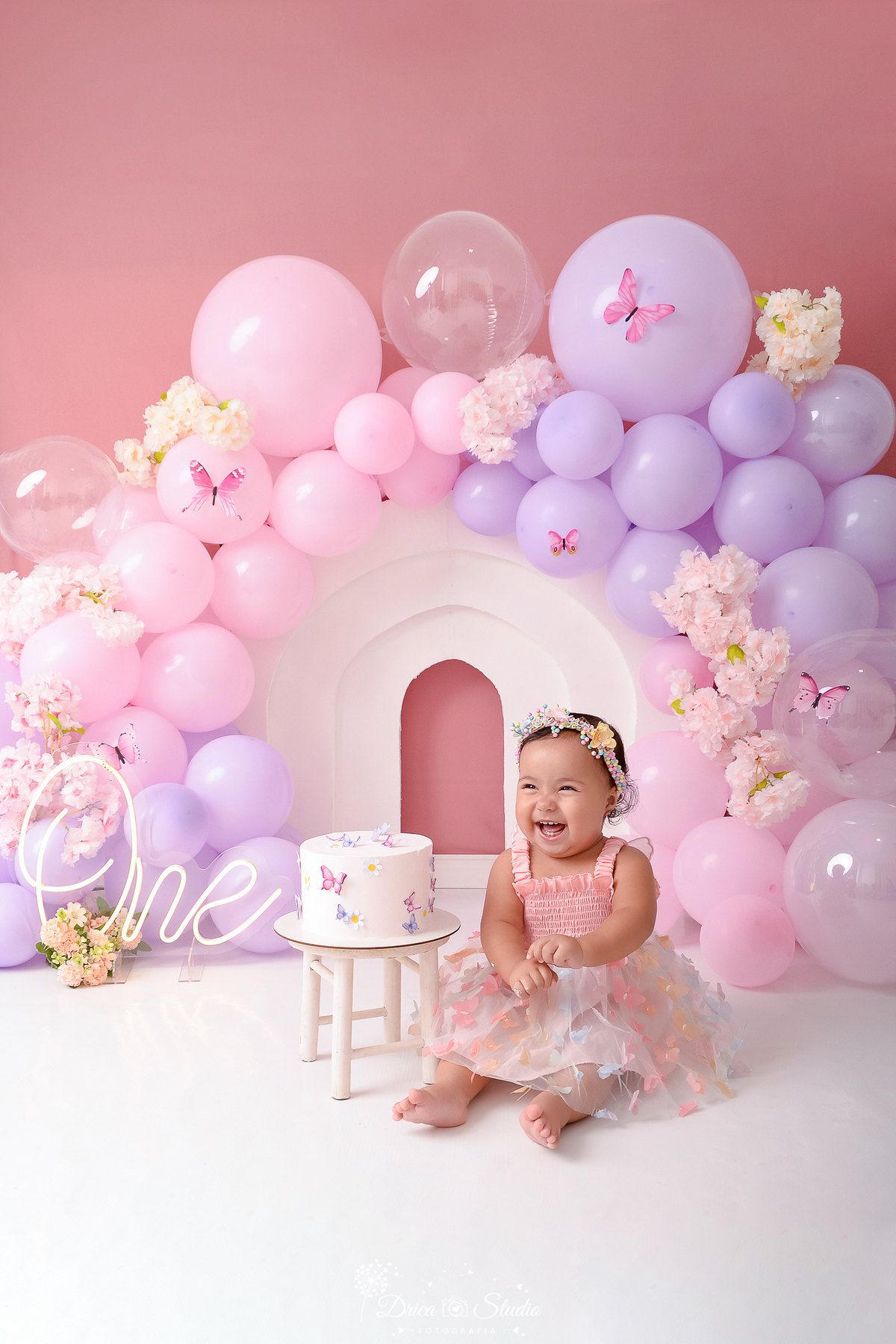 drica studio - ensaio fotográfico infantil - smash the cake - tema jardim de borboletas - xinguara - pará - balões lilas e rosa - borboleta e flores rosa - bolo branco - one de led - arco branco - bebê sorridente usando vestido rosa e tiara com flores 