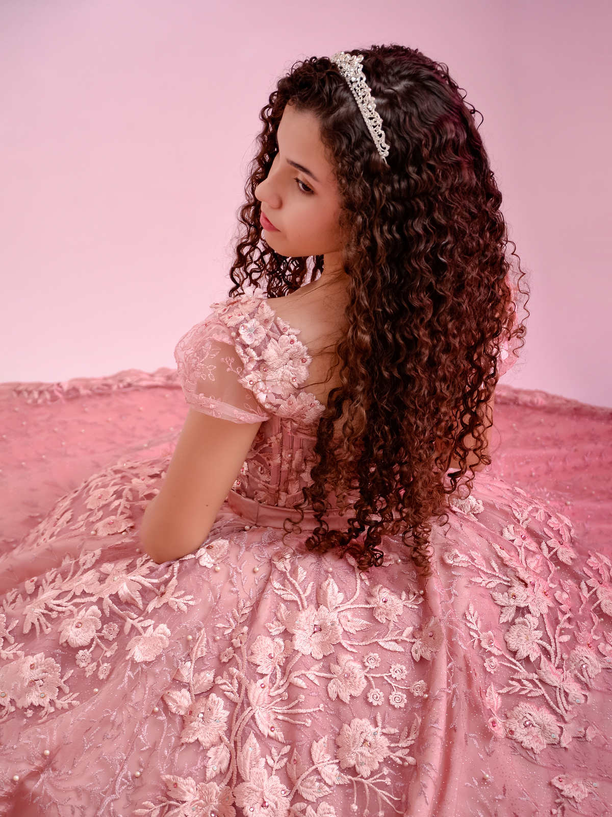 drica studio - ensaio fotografico - debutante 15 anos - aniversário - menina com cabelo cacheado longo usando vestido de festa rosa com renda floral e aplicação de pérolas - debutante sentada no chão de perfil - fundo rose