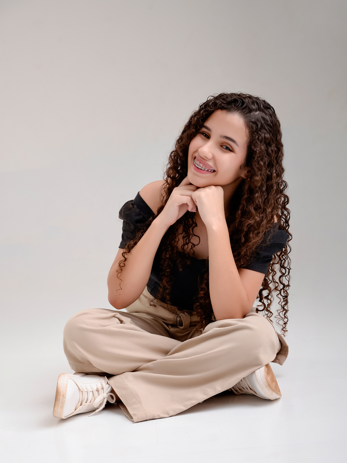 drica studio - ensaio fotografico - debutante 15 anos - aniversário - menina sentada no chão com pés cruzados - blusa preta e calca bege - fundo neutro