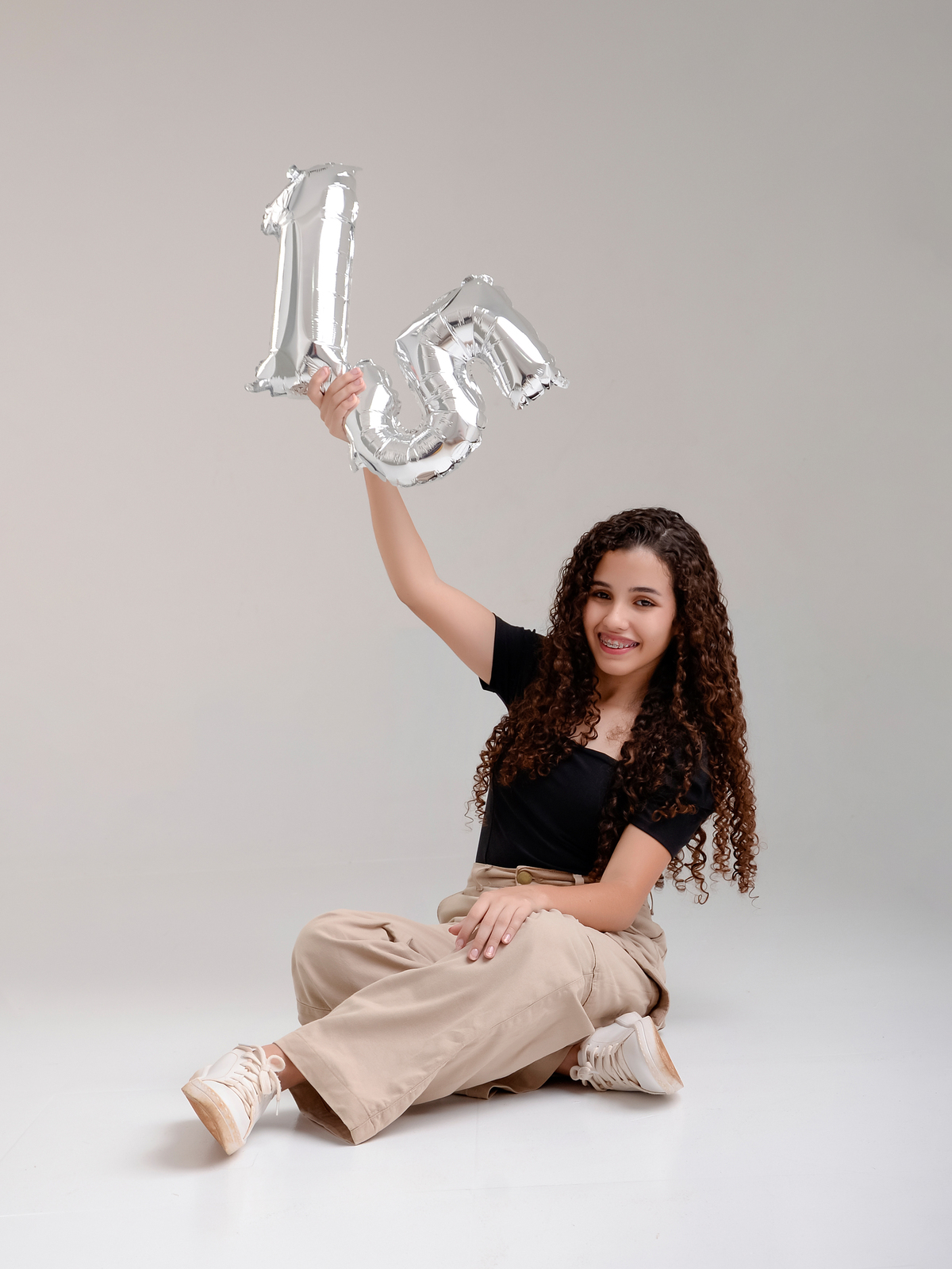 drica studio - ensaio fotografico - debutante 15 anos - aniversário - menina sentada no chão com braço erguido segurando número 15 - blusa preta e calça bege - fundo neutro