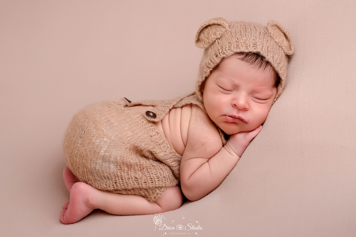 drica studio - ensaio fotografico infantil - newborn masculino - recém nascido dormindo de bruços sobre superficie macia, usando macaquinho de trico bege escuro e goro com orelha de urso- fundo neutro