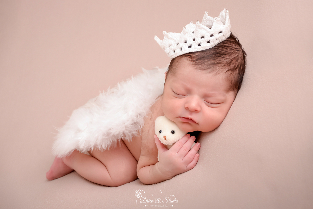 drica studio - ensaio fotografico infantil - recém-nascido - newborn masculino - bebê dormindo de bruços, segurando ursinho branco, asas de anjo sobre suas costa e coroa de crochê sobre sua cabeça - fundo neutro