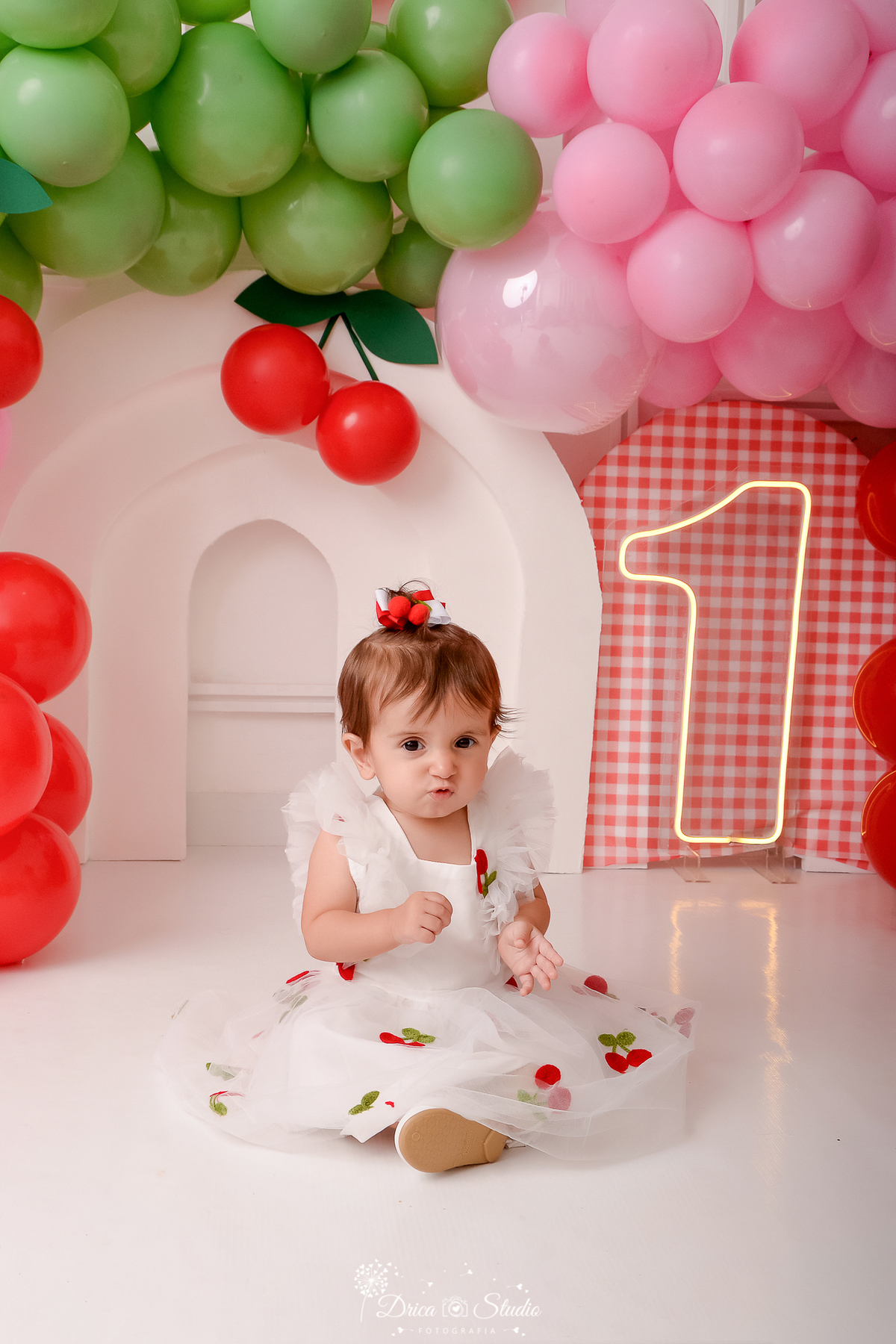 drica studio - ensaio fotografico infantil - smash the cake cerejinha - balões vermelho, painel xadrez com vermelho - arco 3d -1 de led - arco 3d branco - bebê sentada no chão usando vestido branco com tule e apliques de cerejinhas.