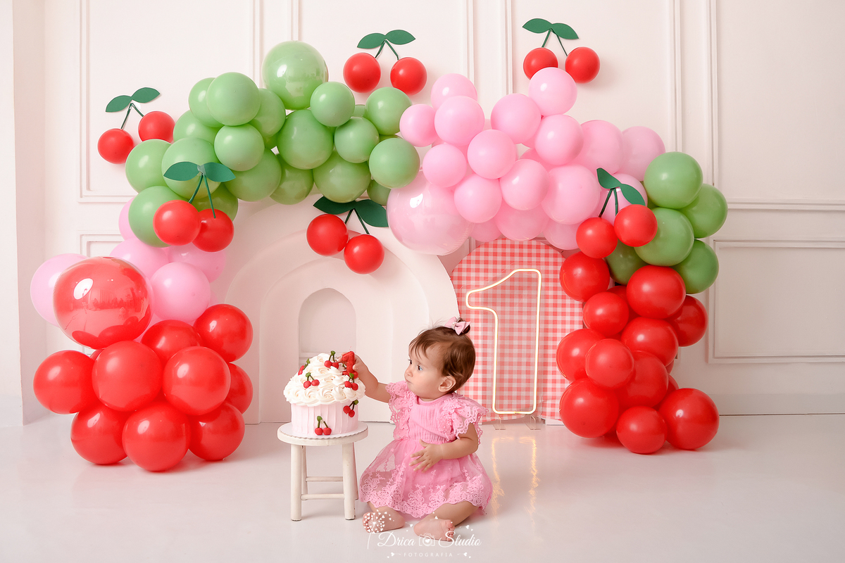 drica studio - ensaio fotografico infantil - smash the cake cerejinha -  -balões vermelho,rosa e verde - cerejinhas de balão - painel xadrez vermelho com branco - bebê sentada no chão pegando bolo, que esta sobre banquinho - fundo neutro