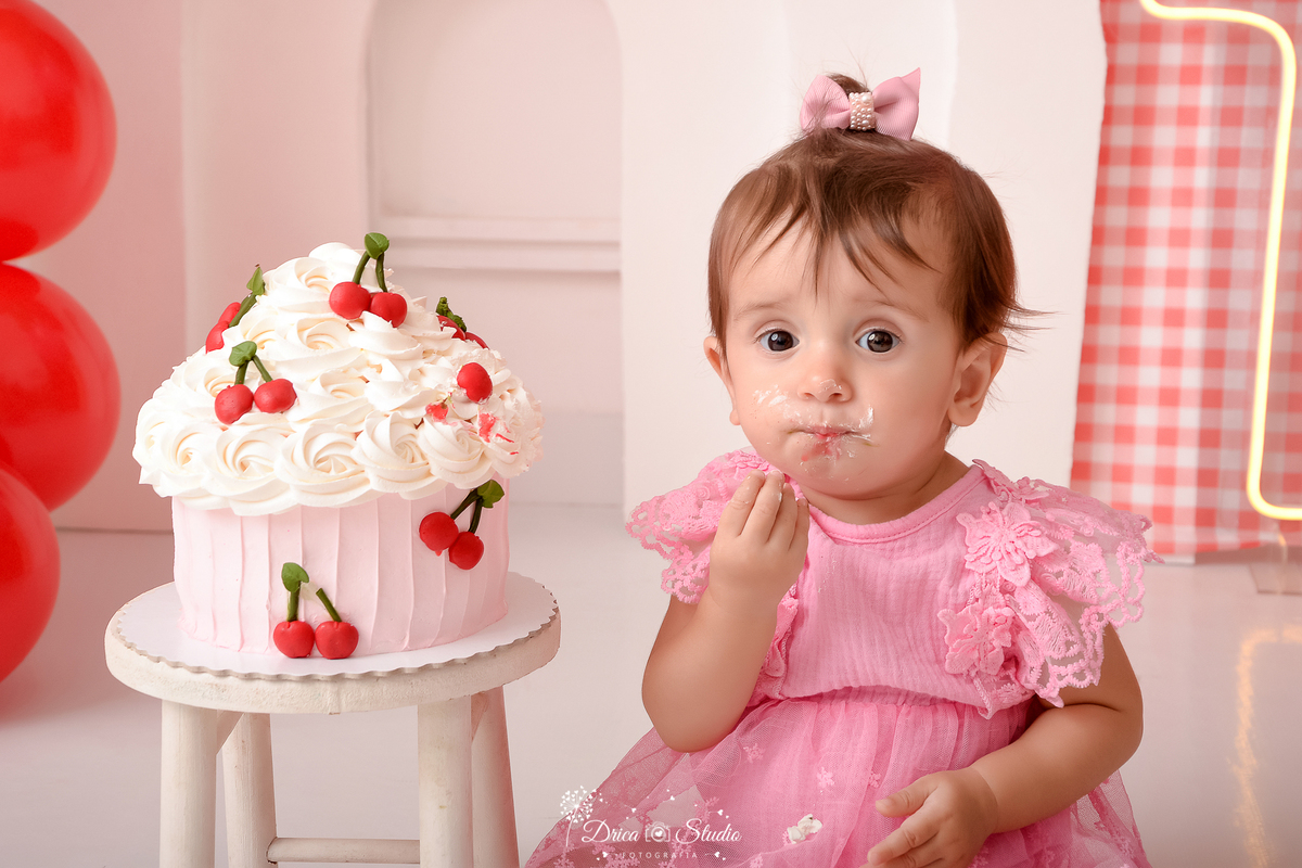 drica studio - ensaio fotografico infantil - smash the cake cerejinha - bebê sentada no chão comendo bolo- vestido rosa com renda - cupcake branco com cerejinhas - arco 3d branco - painel xadrez vermelho com branco - balões vermelho - fundo branco