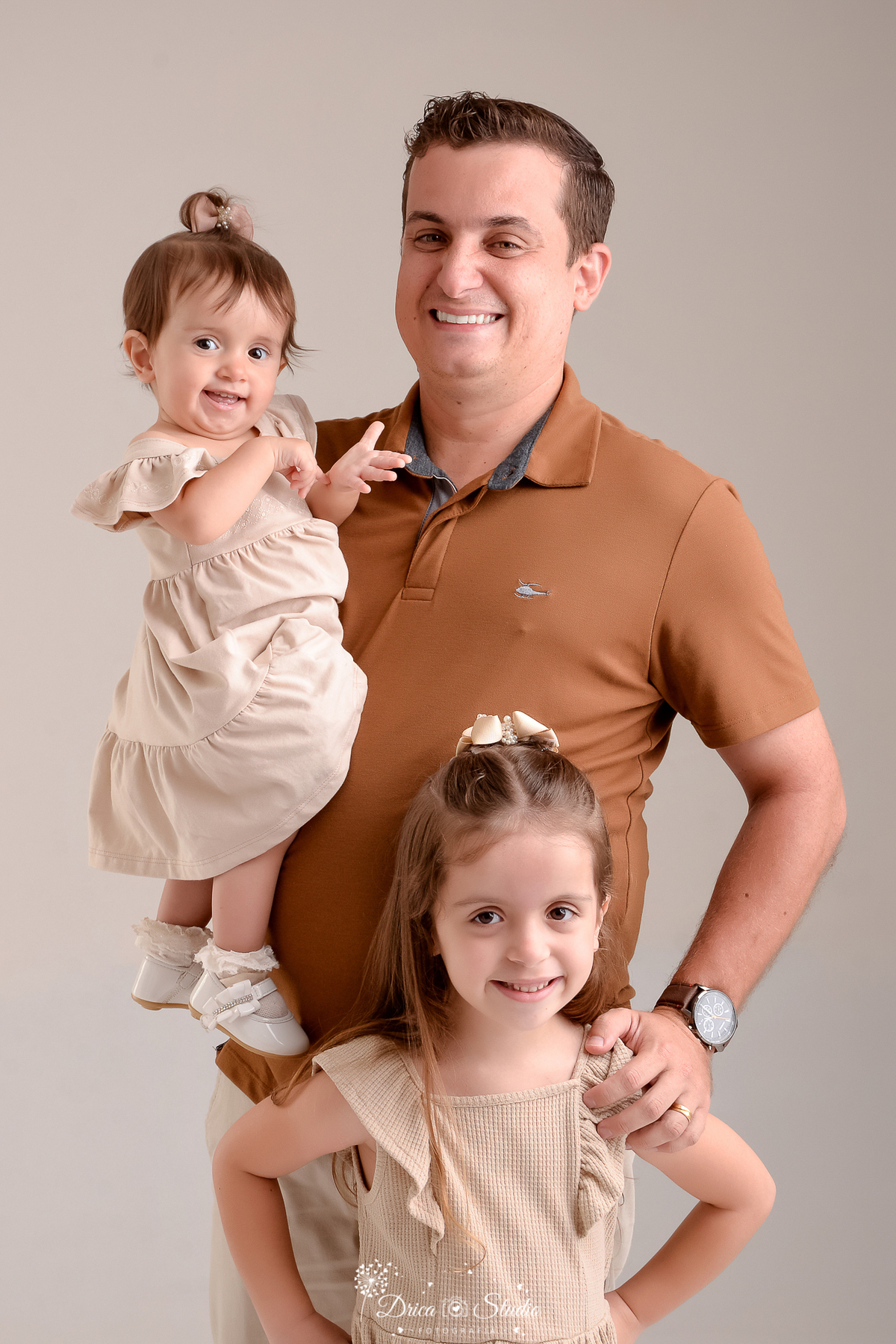 drica studio - ensaio fotografico em familia - xinguara - pará - pai posando para foto com as filhas, no colo a bebê e a sua frente a filha mais velha - as meninas usam vestido bege claro e o pai camiseta marrom e calça bege- fundo neutro