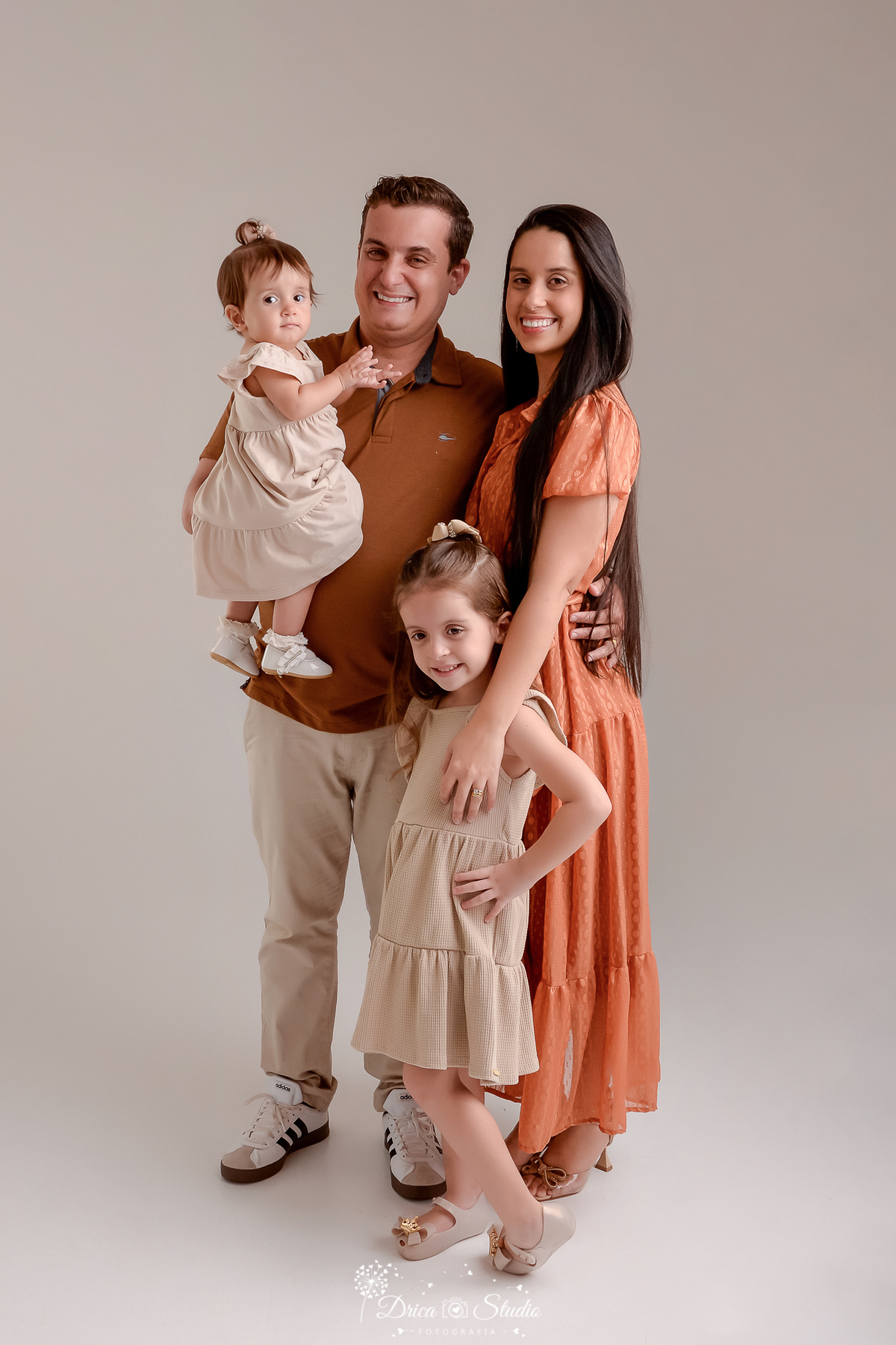 drica studio - ensaio fotografico em familia - xinguara - pará - casal abraçado - pai segurando filha no colo e filha mais velha a frente da mãe - meninas usam vestido bege claro - mae vestido cor telha e pai camiseta marrom e calça bege - fundo neutro