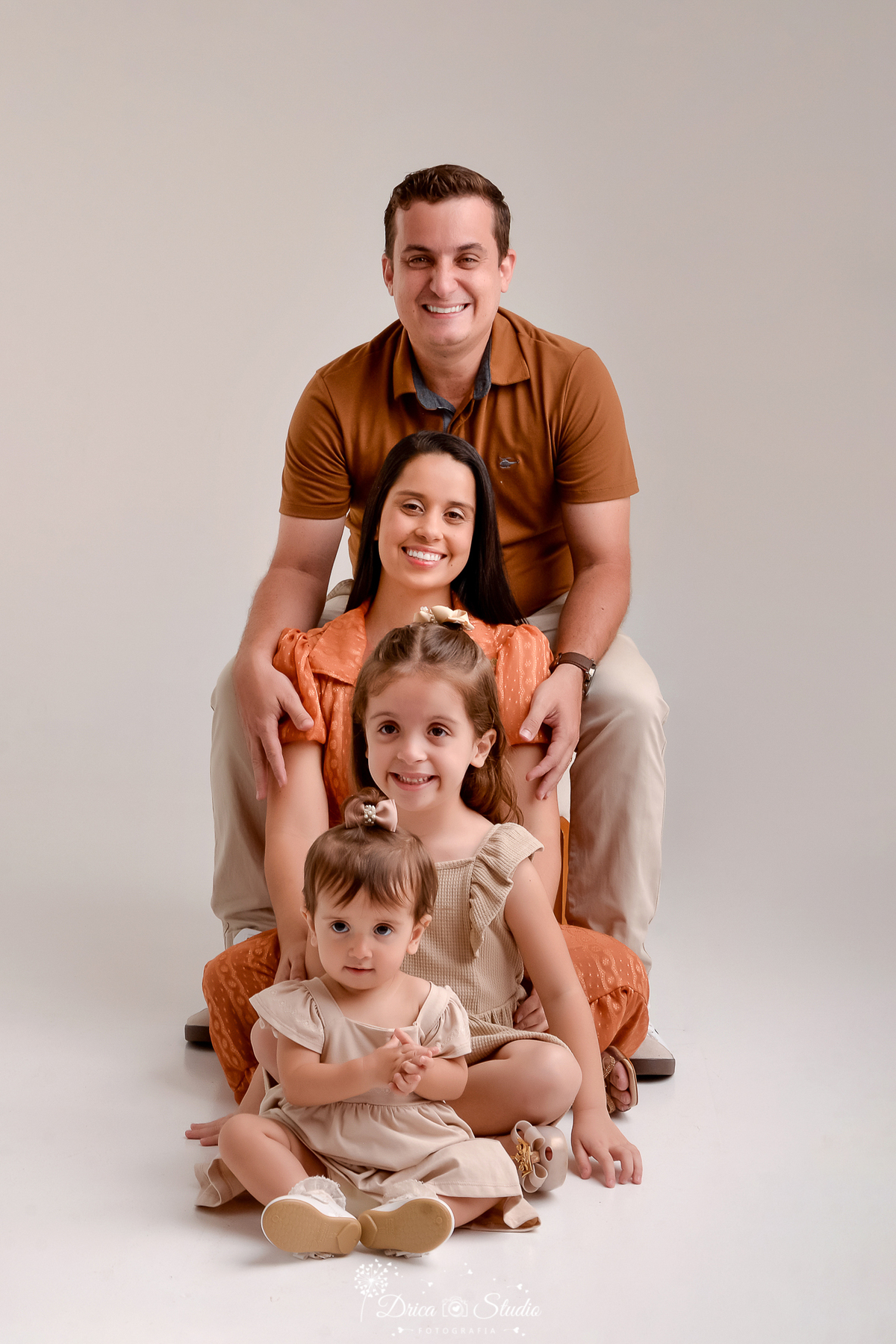 drica studio - ensaio fotografico em familia - xinguara - pará - familia posa um a frente do outro sentado - meninas vestem vestido bege, mãe vestido cor telha e pai camiseta marrom e calça bege - fundo neutro