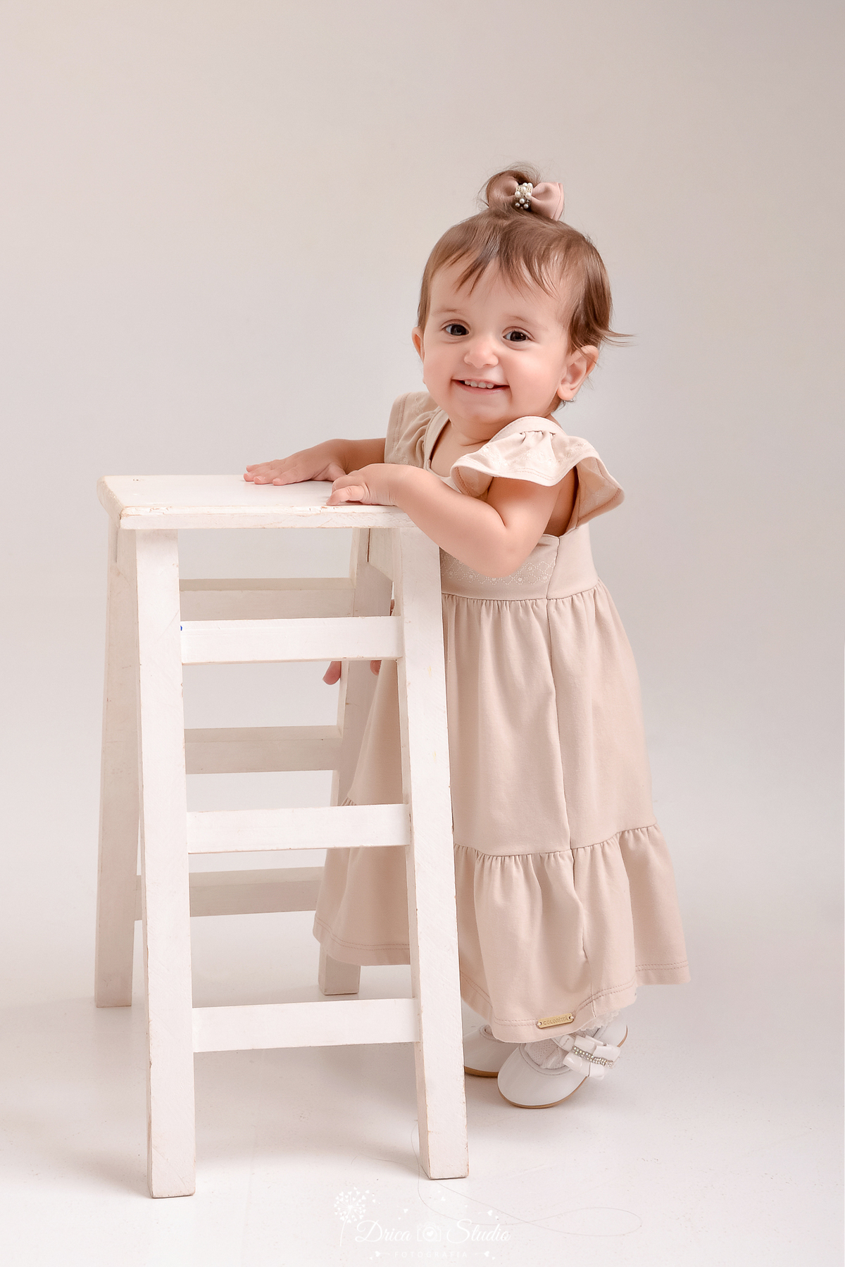 drica studio - ensaio fotografico em familia - xinguara - pará - bebê em pé se apoiando em banquinho de branco - vestido bege claro - fundo neutro