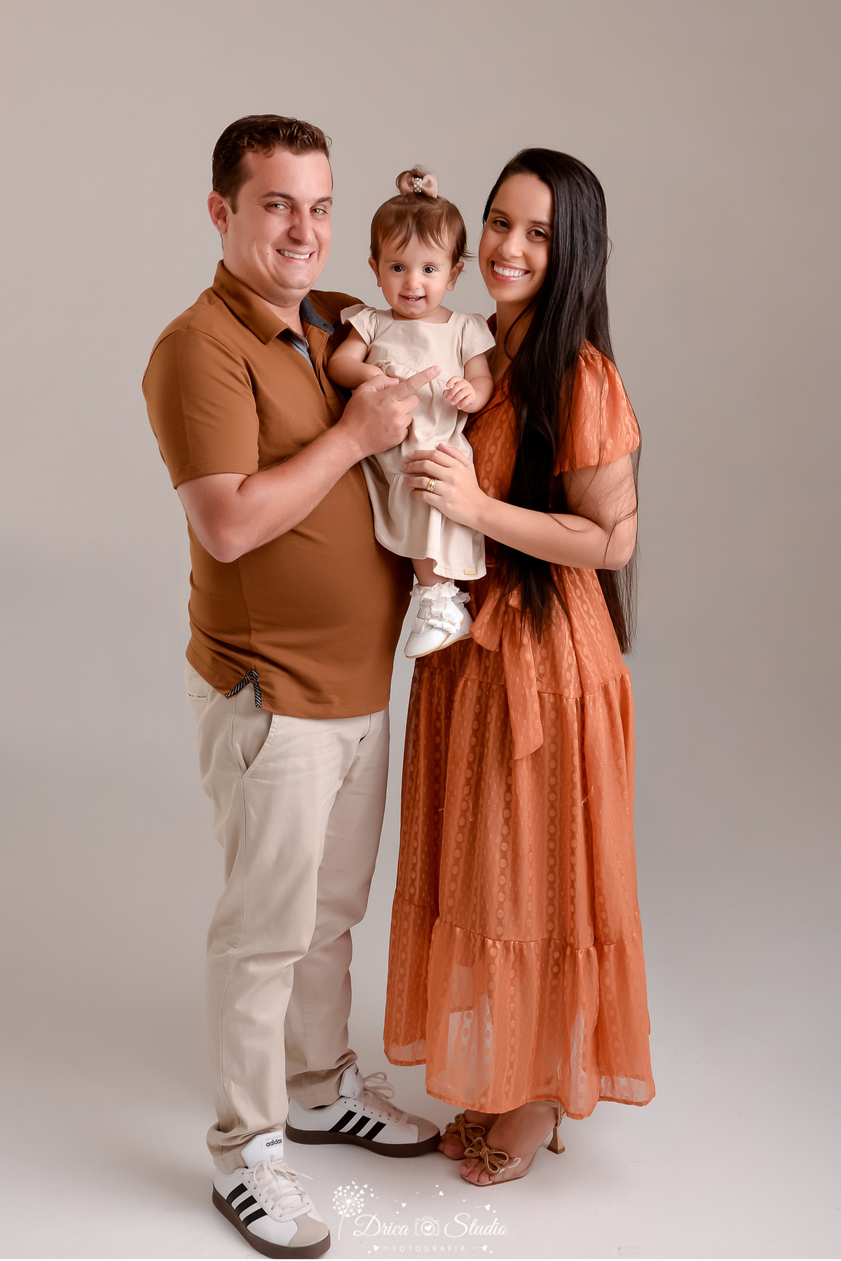 drica studio - ensaio fotografico em familia - xinguara - pará - casal segura bebê no colo - mulher usa vestido cor telha - pai camisa marrom e calça bege clara e a bebê vestido bege claro - fundo neutro