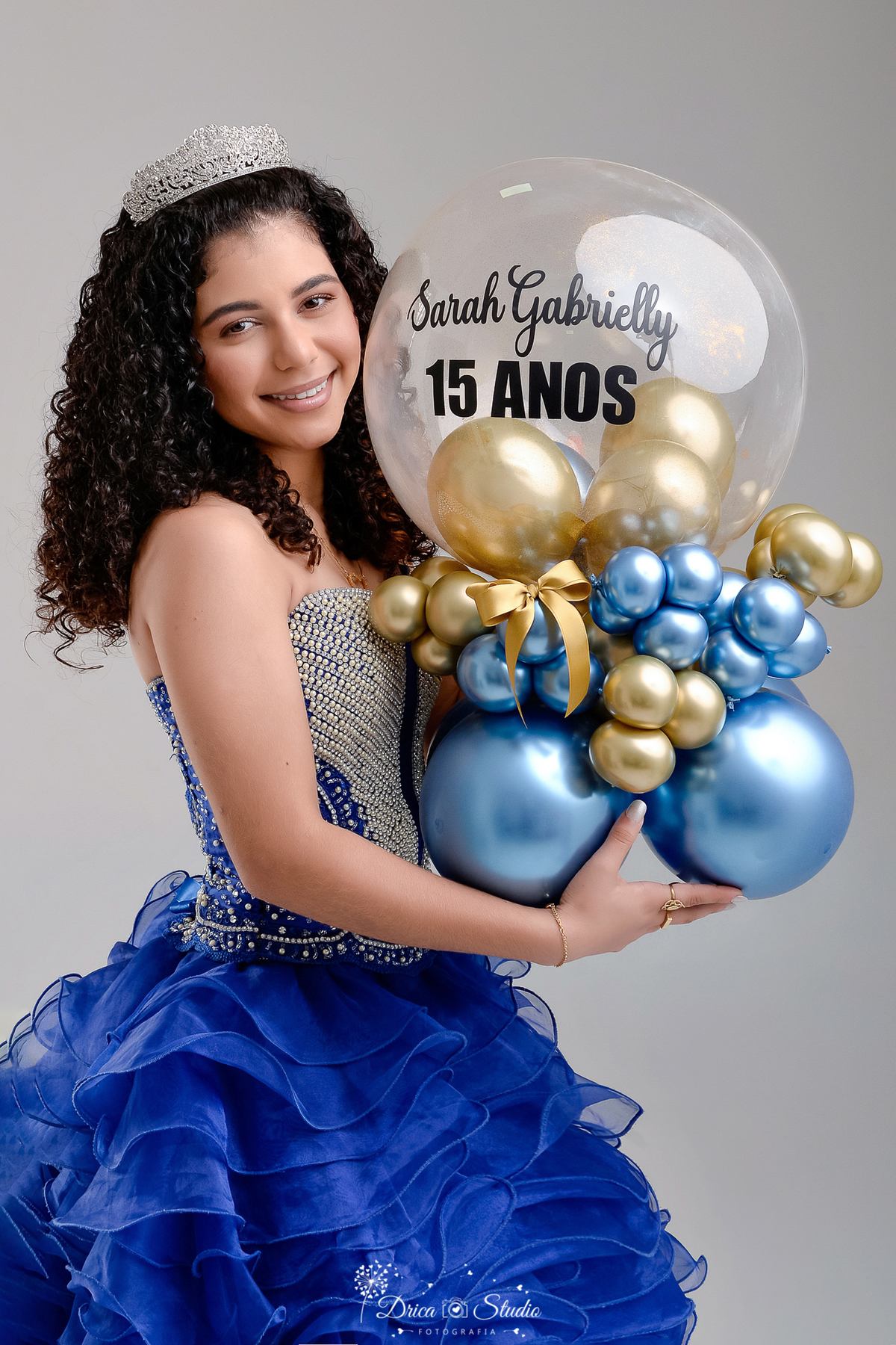 rica studio - ensaio fotografico - 15 anos - debutante - xinguara - pará  - jovem segurando cacho de balões dourado, azul e boboo transparente com a frase 15 anos - vestido azul com pedraria - fundo neutro