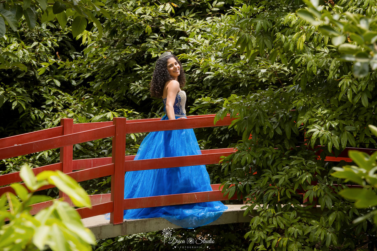 drica studio - ensaio fotografico - 15 anos - debutante - xinguara - pará  - ensaio ao ar livre - jovem usando vestido estilo princesa azul, posa para foto em passarela em meio ao jardim tropical