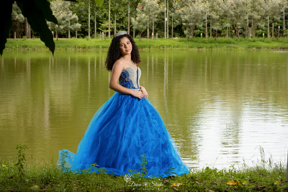 drica studio - ensaio fotografico - 15 anos - debutante - xinguara - pará -ensaio ao ar livre - joves debutante usando vestido de festa azul, com pedraria e delicada cora, posa para foto em frente a logo em jardim tropical