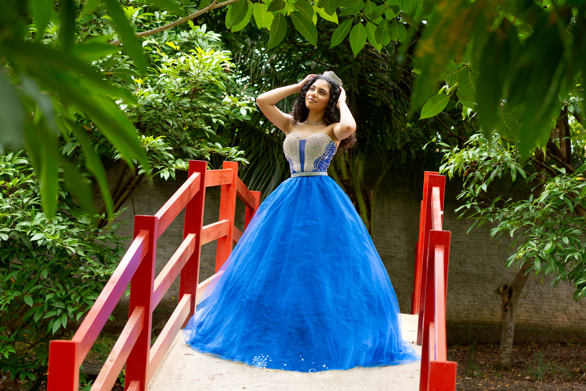 drica studio - ensaio fotografico - 15 anos - debutante - xinguara - pará-  - ensaio ao ar livre - debutante usando vestido azul de festa, posa para foto segurando delicada corao em sua cabeça, em meio a passarela com corrimão vermelho -  em meio a bosque