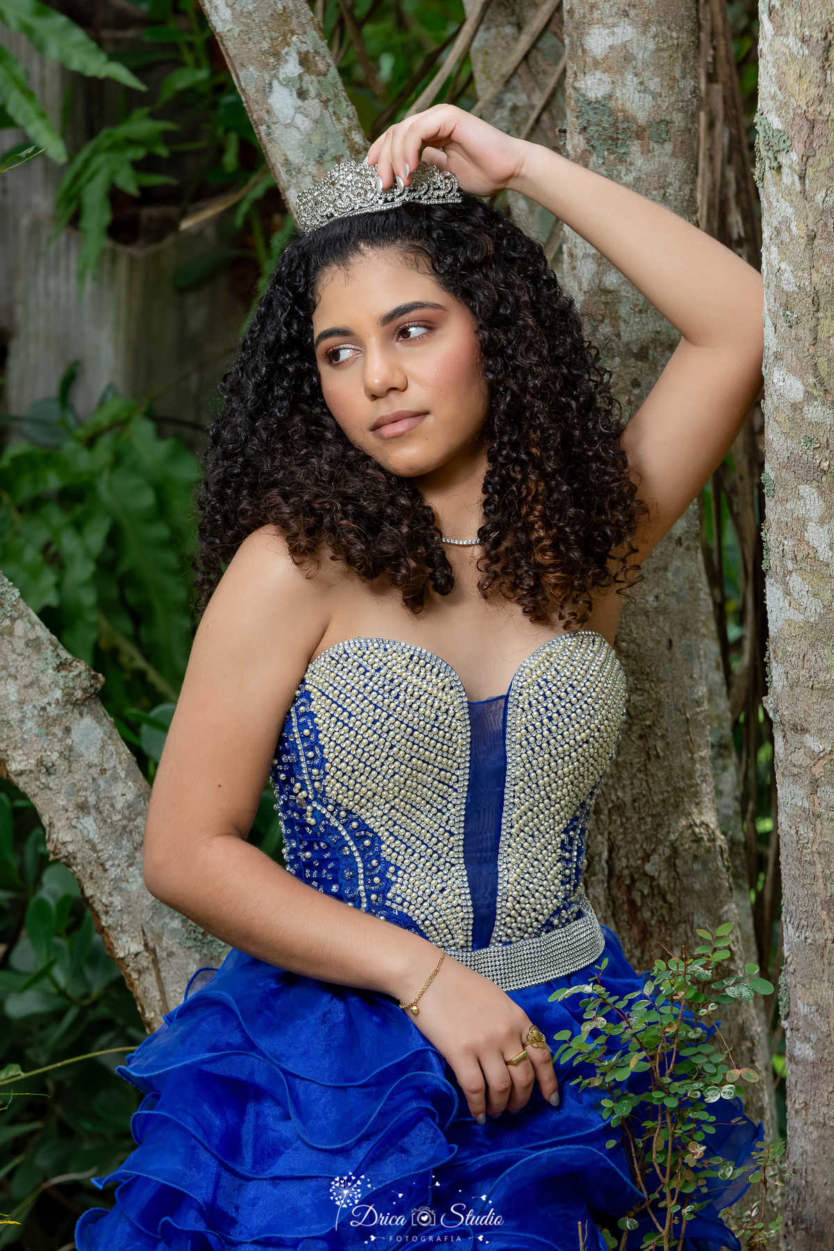 drica studio - ensaio fotografico - 15 anos - debutante - xinguara - pará - ao ar livre - vestido de festa azul, corpo com pedraria e saia com muitos babado - ao fundo um bosque - jovem com braço erguido encostado em uma arvore