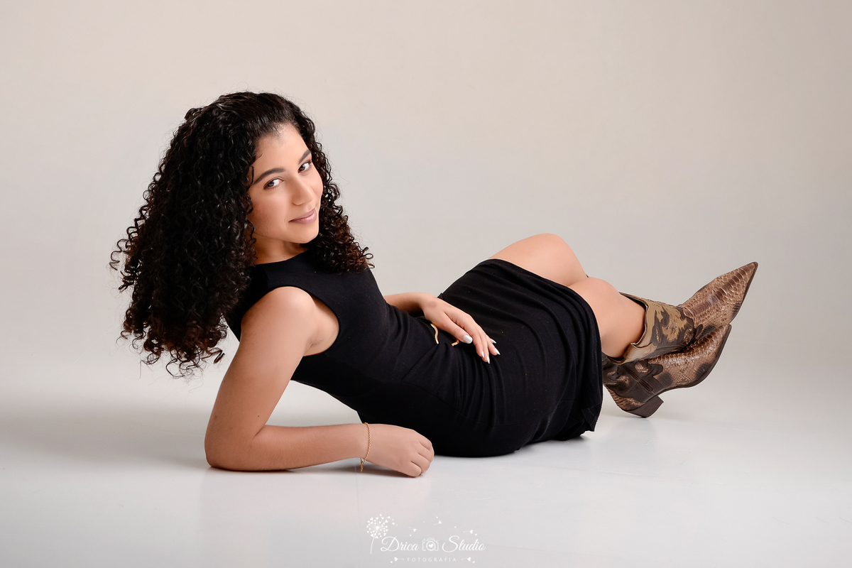 drica studio - ensaio fotografico - 15 anos - debutante - xinguara - pará - jovem deitada de lado em um estúdio com fundo neutro claro - vestido preto - bota cano longo  marrrom - fundo neutro