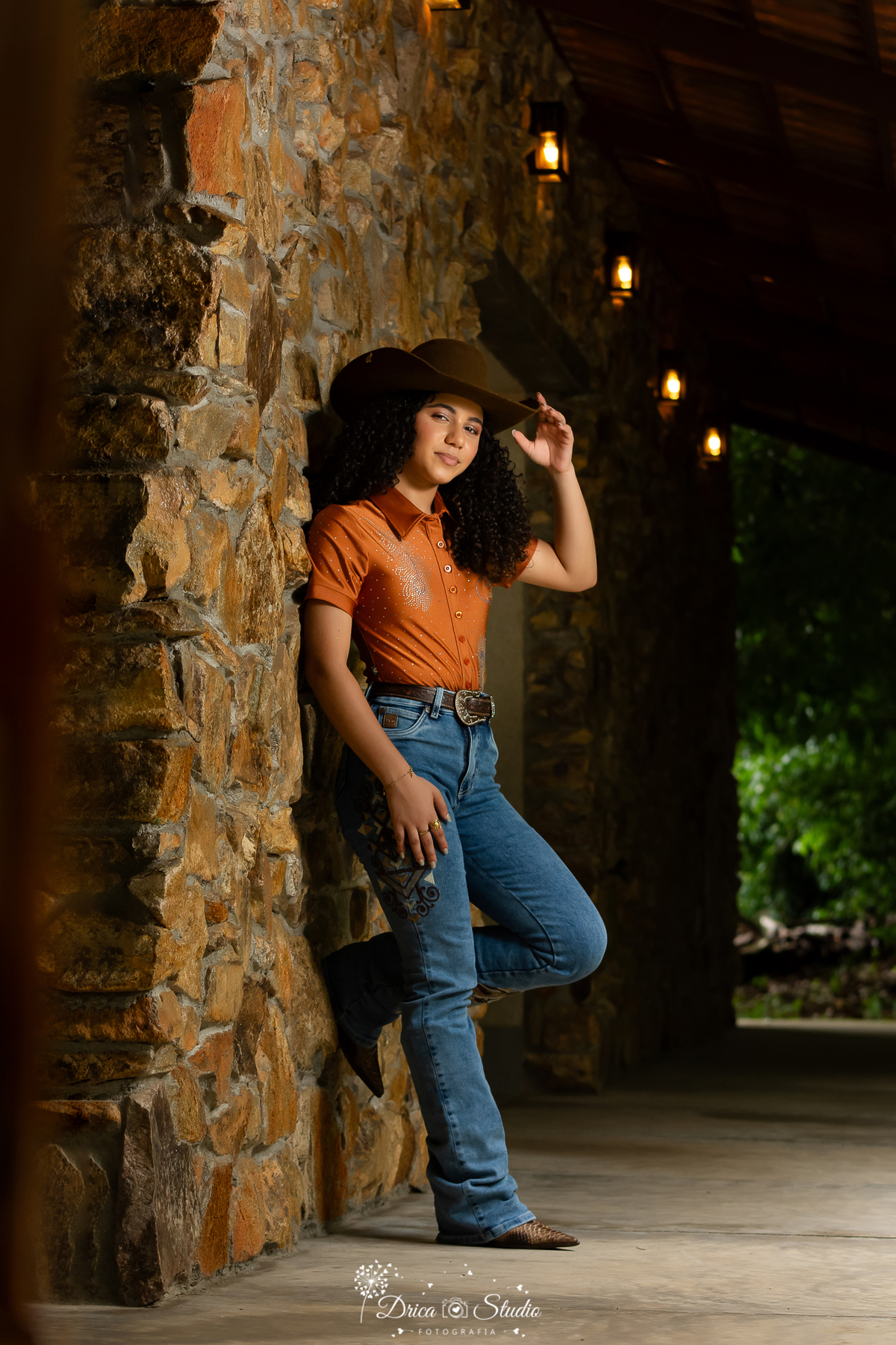 drica studio - ensaio fotografico - 15 anos - debutante - xinguara - pará - jovem usando calça jeans com bordados na perna e camisa terracota com detalhes em bordado - chapeú marrom- bota cano longo estilo pela de cobra marrom - parede de pedras rusticas