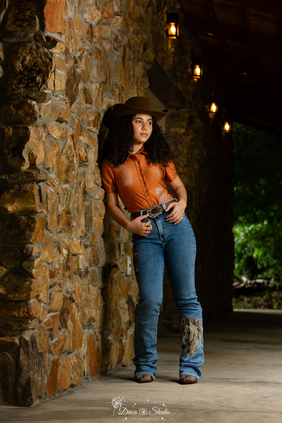 drica studio - ensaio fotografico - 15 anos - debutante - xinguara - pará - jovem posa para foto encostada em parece de pedras rusticas- ela usa calça jeans e camisa na cor terracota com detalhes em bordado - chapeú, cinto e bota marrom - 