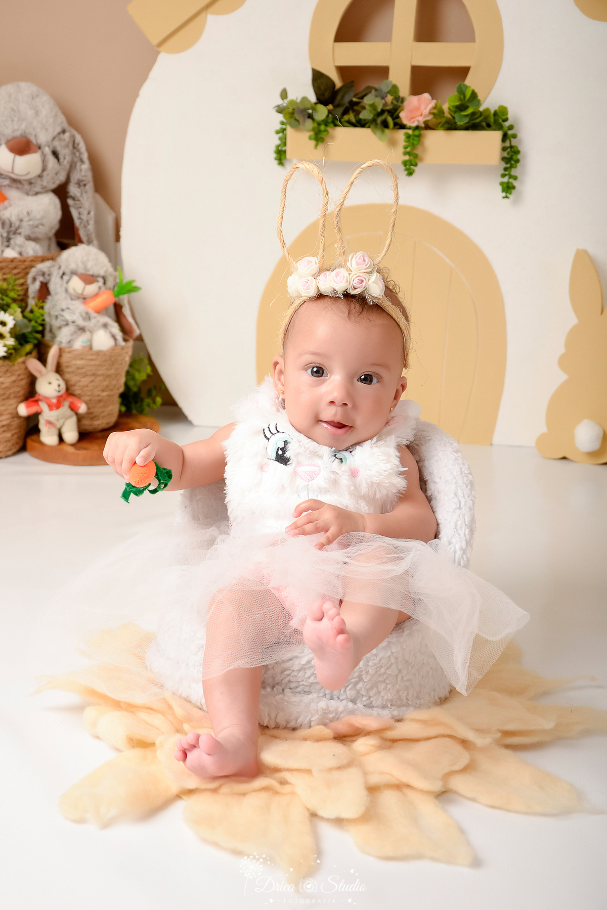 drica studio - ensaio fotografico - acompanhamento mensal - ensaio infantil - páscoa - xinguara - pará - bebê sentada em poltrona branca, vesti vestido branco de tule- cestos de palha - coelhos de pelúcia - tiara de flores - casinha banca com porta bege -
