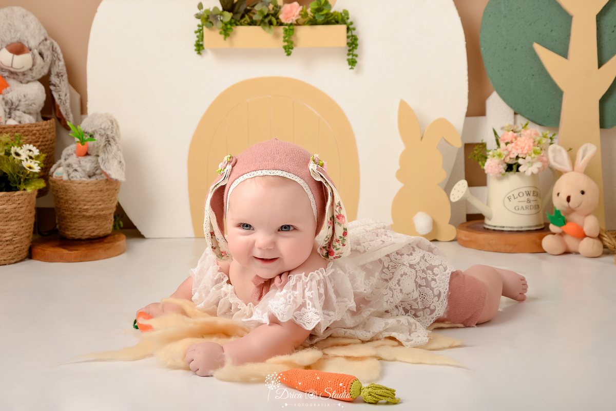 drica studio - ensaio fotografico - acompanhamento mensal - ensaio infantil - páscoa - xinguara - pará -bebê deitada de bruços sobre tapete felpudo - ela veste bata de renda e touquinha com orelhas de coelho - cenoura de pelucia - cestos de palha-regador