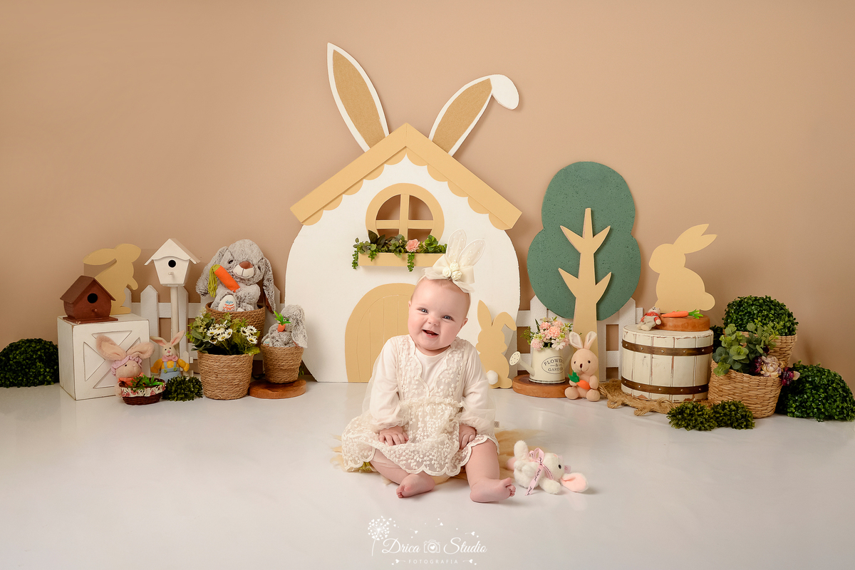 drica studio - ensaio fotografico - acompanhamento mensal - ensaio infantil páscoa - xinguara - pará - bebê sentada no chão, usnado vestido de renda e tiara de flores - casinha de coelho - coelhos de pelucia - arvore de isopor - balde branco - regador 
