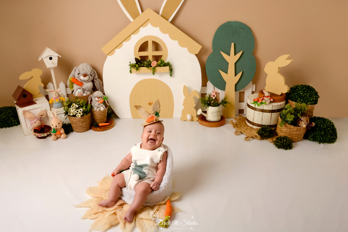 drica studio - ensaio fotografico - acompanhamento mensal - ensaio infantil páscoa - xinguara - pará - bebê sentado em poltrona branca -  usando chapeú de palha com orelhas de coelho - cestos de palhas com flores - regador com flores - coelhos de pelucia 