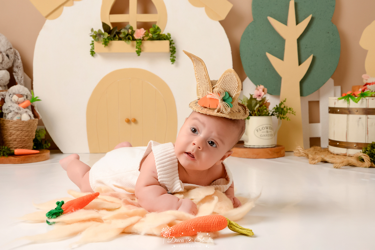 drica studio - ensaio fotografico - acompanhamento mensal - ensaio infantil páscoa - xinguara - pará - bebê deitado no chão sobre tapete felpudo - macaquinho bege de trico e chapeú de palha com orelhas de coelho - casa de coelho - arvore de isopor - balde