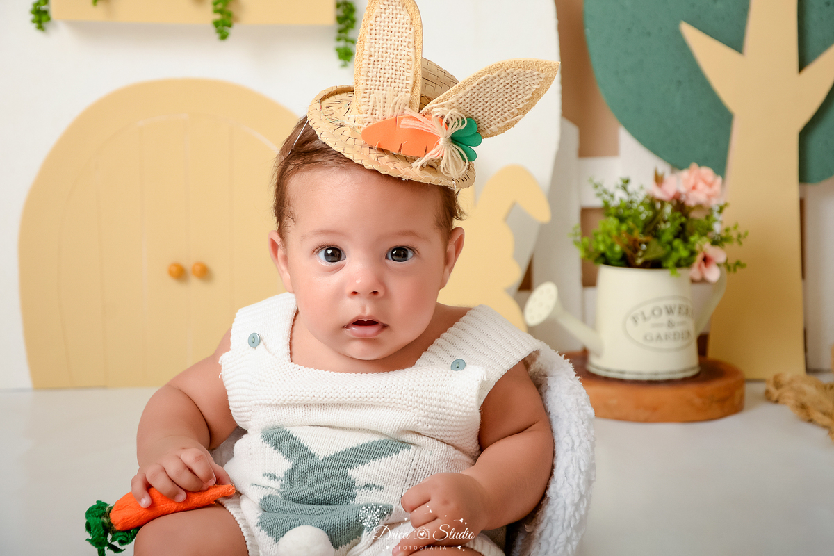 drica studio - ensaio fotografico - acompanhamento mensal - ensaio infantil páscoa - xinguara - pará - bebê sentado em poltrona, usando bory de tricou branco com chapeú com orelhas de coelho - casinha branca com detalhes bege - regador branco com flores