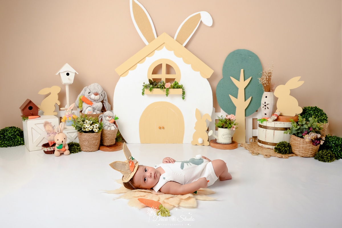 drica studio - ensaio fotografico - acompanhamento mensal - ensaio infantil páscoa - xinguara - pará - bebê deitado no chã- chapeú com orelhas de coelho - casinha coelho branca - coelhos de pelúcia - cestos de palha- arbustos - caixotes branco 