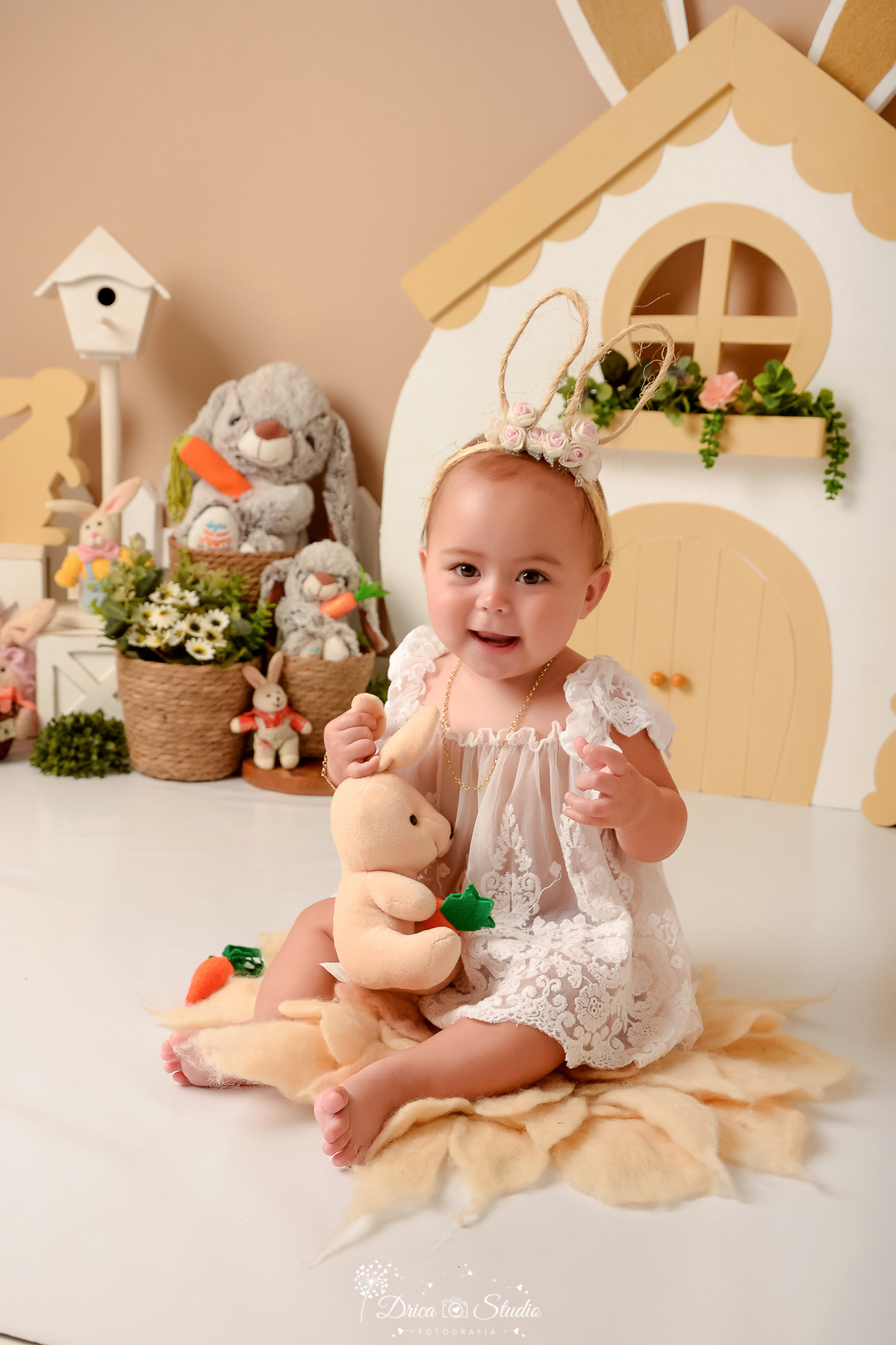 drica studio - ensaio fotografico - acompanhamento mensal - ensaio infantil páscoa - xinguara - pará - bebê sentada no chão, segurando coelho de pelucia, usa batinha de renda e tiara com orelhas de coelho - cesto de palha - coelhos de pelúcia - casinha