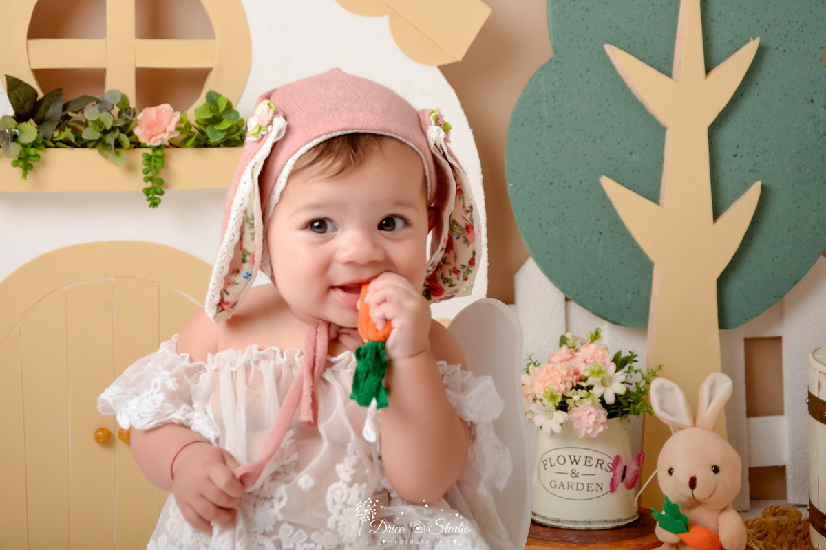 drica studio - ensaio fotografico - acompanhamento mensal - ensaio infantil páscoa - xinguara - pará - bebê mordendo cenora de pano - regador com flores - batinha branca - toca com orelhas de coelho - árvore de isopor - casa branca - ceolho de pelucia
