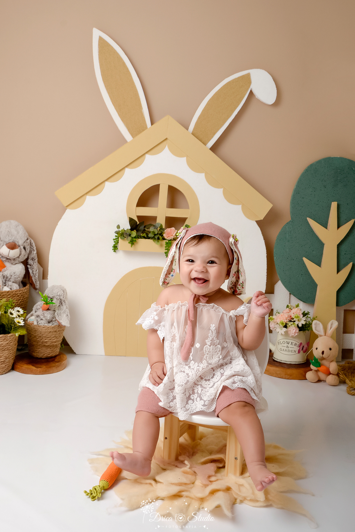 drica studio - ensaio fotografico - acompanhamento mensal - ensaio infantil páscoa - xinguara - pará - bebê sentada no chão - usando bata de renda e toca com orelhas de coelho - caa branca - árvore de isopor - cestos de palha - regador com flores -coelhos