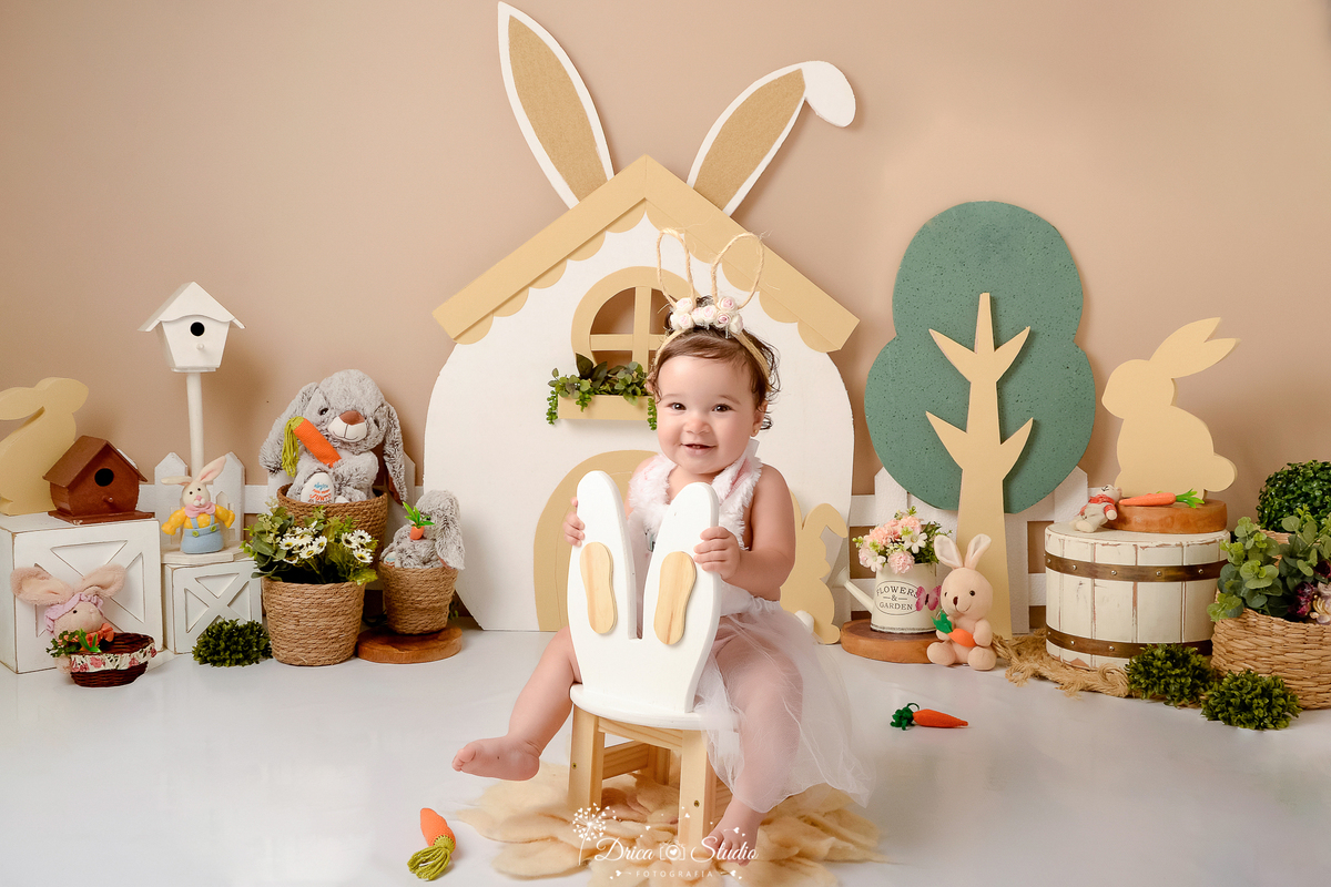 drica studio - ensaio fotografico - acompanhamento mensal - ensaio infantil páscoa - xinguara - pará - bebê sentada em banquinho de orelhas de coelho - casa branco-coelhos de pelúcia-cesto de palha-balde branco-regador com flores- fundo neutro