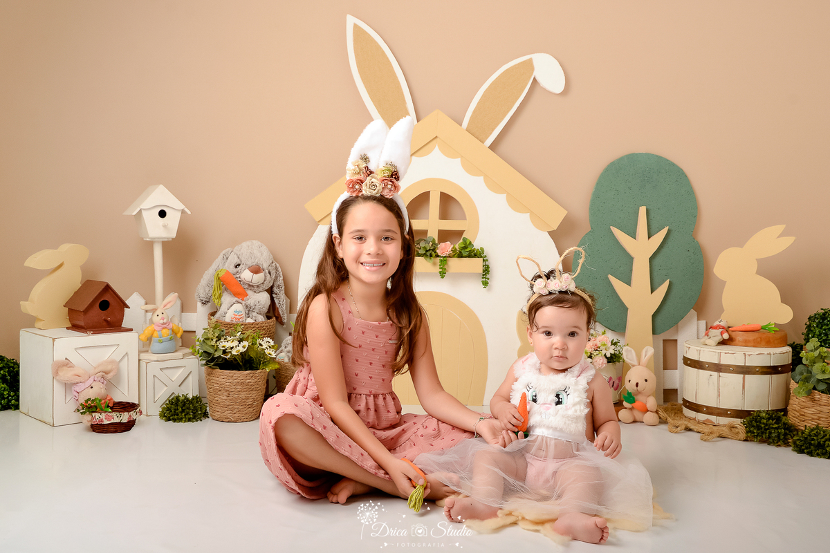drica studio - ensaio fotografico - acompanhamento mensal - ensaio infantil páscoa - xinguara - pará - bebê sentada no chão ao lado da irmã mais velha, ambas usa orelhinhas de coelho - coelhos de pelúcia - casa de coelho - balde branco - cesto de palha - 