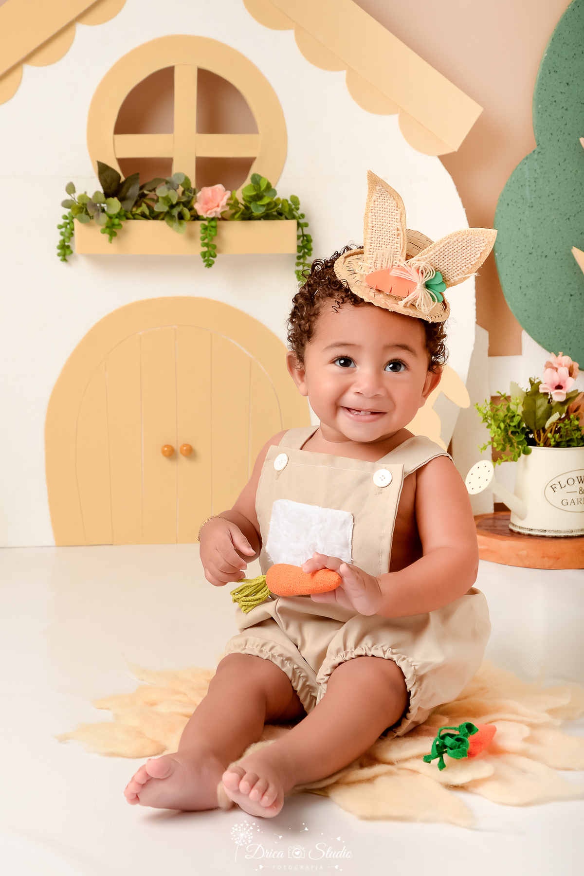drica studio - ensaio fotografico - acompanhamento mensal - ensaio infantil páscoa - xinguara - pará - bebê sentado no chão vestido com macaquinho bege e chapeú de palha com orelhas de coelho  - casa branca - regador branco com flores - cenoura de tecido