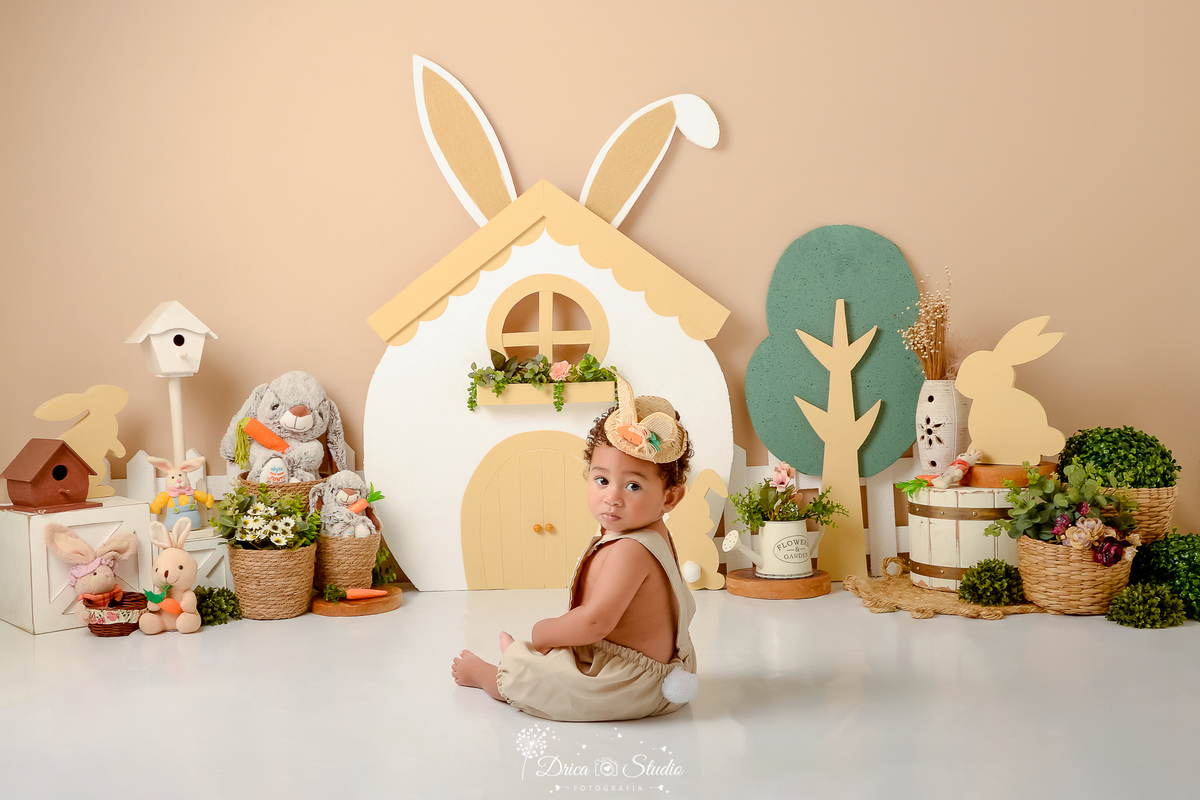 drica studio - ensaio fotografico - acompanhamento mensal - ensaio infantil páscoa - xinguara - pará - bebe sentado no chão com macaquinho bege e chapeú de com orelhas de coelho - casa de coelho - casa de passarinho - balde branco - coelhos de pelucia -