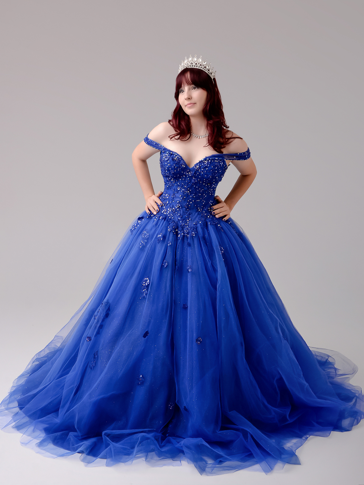 drica studio - fotos 15 anos - aniversario - debutante - vestido azul - estúdio - foto de longe - com as mãos na cintura - usando coroa -  sorrindo - fundo na cor cinza 