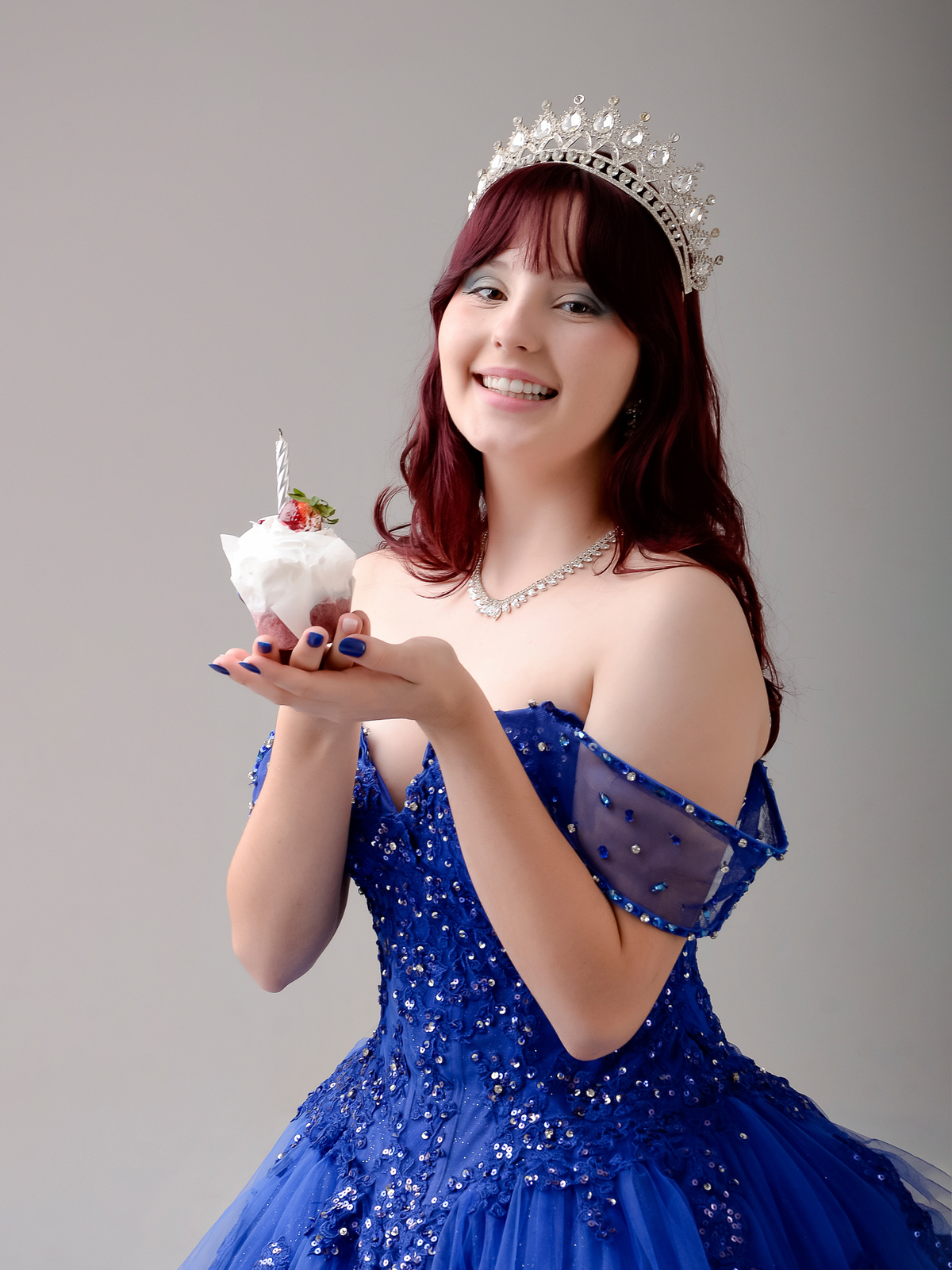 drica studio - fotos 15 anos - aniversario - debutante -  vestido azul - estúdio - foto sorrindo -  segurando cupcake com vela - usando coroa -  fundo na cor cinza 