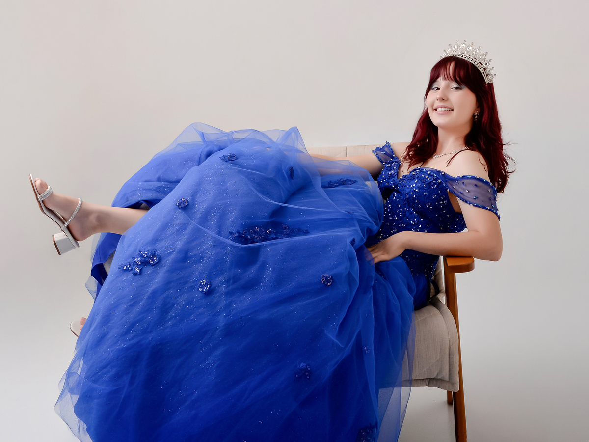 drica studio - fotos 15 anos - aniversario - debutante - vestido azul - sentada numa poltrona - sorrindo - usando coroa - estúdio - fundo na cor cinca  