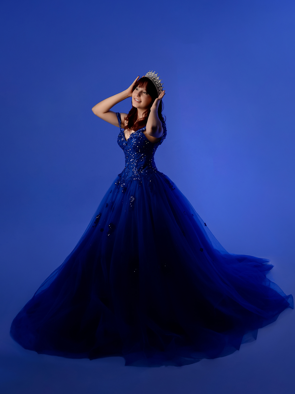 drica studio - fotos 15 anos - aniversario - debutante - estúdio - vestido azul - em pé -  segurando a coroa com as duas mãos - sorrindo - luz de fundo azul escuro 