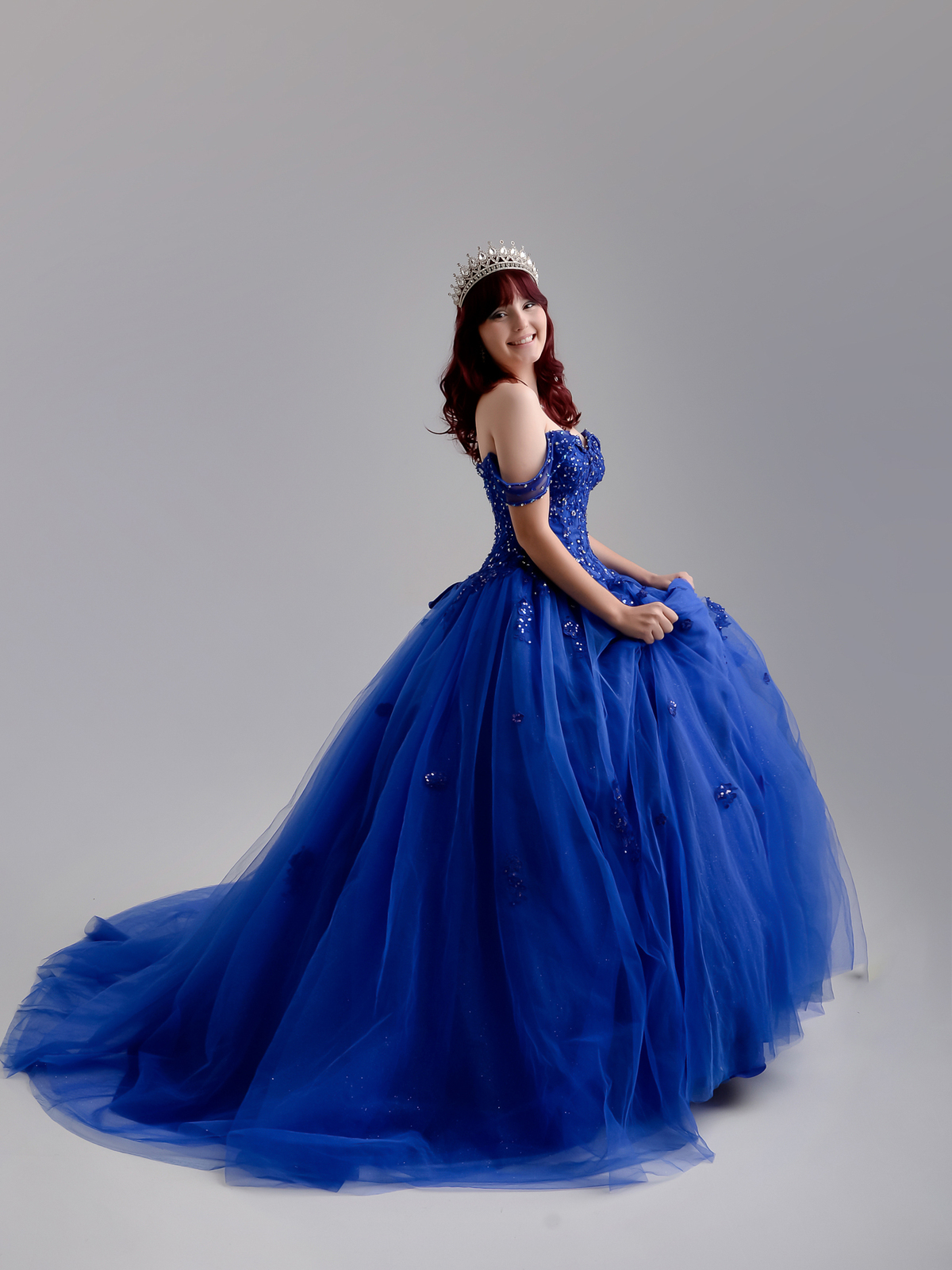 drica studio - ensaio de 15 anos - aniversário - debutante - fotos 
estúdio - foto de perfil - segurando o vestido - usando coroa - sorrindo - vestido azul - fundo na cor cinza 