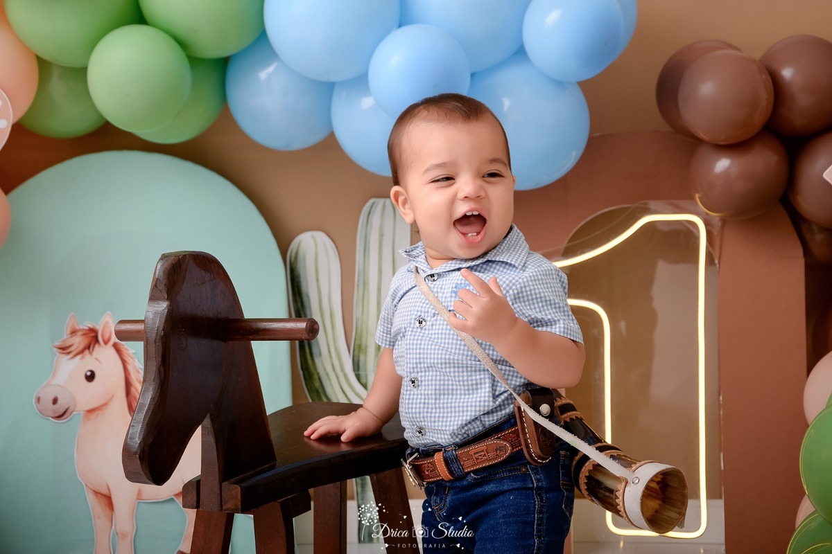 drica studio - ensaio fotografico - fotos em estúdio - xinguara/pá - smash the cake - cenário com balões - cavalos e cacto - 1 ano - bebê em pé - segugrando em um cavalinho de madeira - usando camisa xadrez, calça, cinto e berrante 