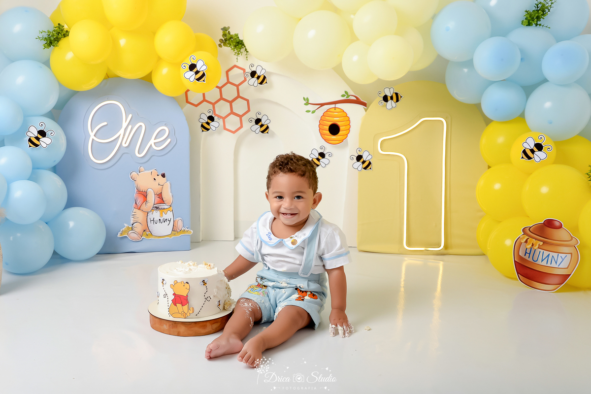 drica studio - smash the cake - 1 ano - aniversario - tema ursinho pooh - cenário com balões - balões azuis e amarelos - abelhas - favo de mel - pote de mel - one de led - 1 de led - bebê sentado - com a mãezinha no bolo - sorrindo 