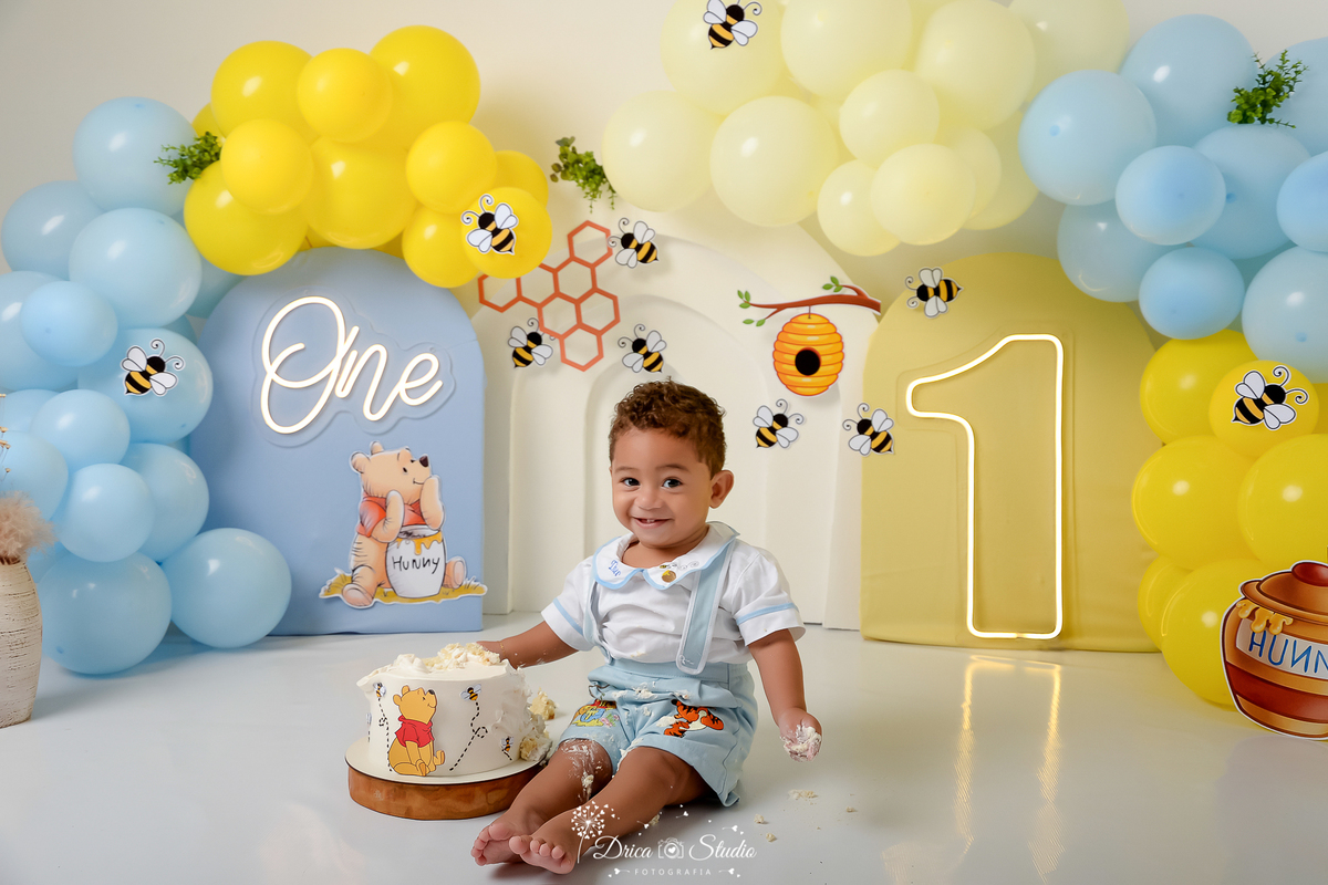 drica studio - smash the cake - 1 ano - aniversario - tema ursinho pooh - cenário com balões - balões azuis - balões amarelos - abelhas - favo de mel - pote de mel - one de led - 1 de led - painel azul -  bebê sentado - brincando com o bolo - sorrindo 