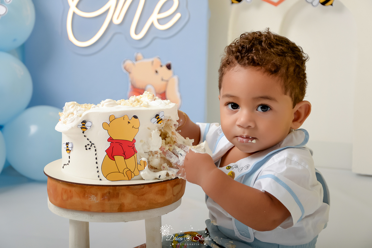 drica studio - smash the cake - 1 ano - aniversario - tema ursinho pooh - one de led - painel azul - painel branco - balões azuis - bebê sentado - tocando o bolo 