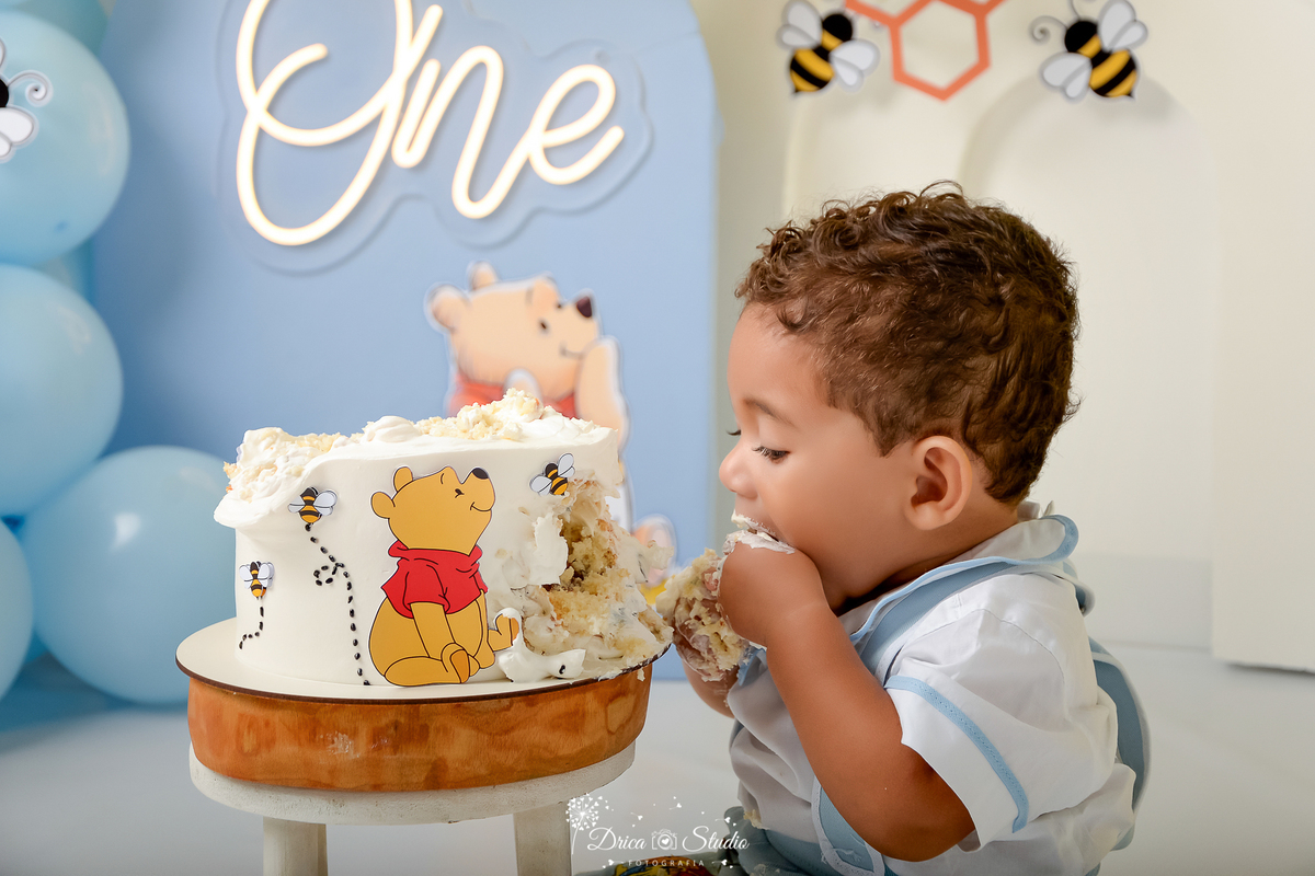 drica studio - smash the cake - 1 ano - aniversario - tema ursinho pooh - ao fundo painel azul e branco - abelhas - favo de mel - balões azuis - bebê comendo bolo 