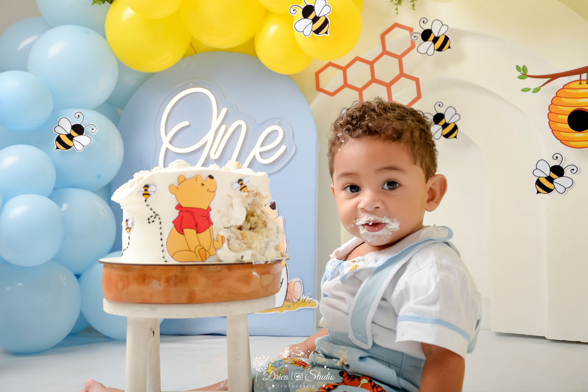 drica studio - smash the cake - 1 ano - aniversario - tema ursinho pooh - cenário com balões - balões azuis - Balões amarelos - abelhas, favo de mel - one de lede - painel azul - bolo do ursinho pooh - bebê comendo bolo  