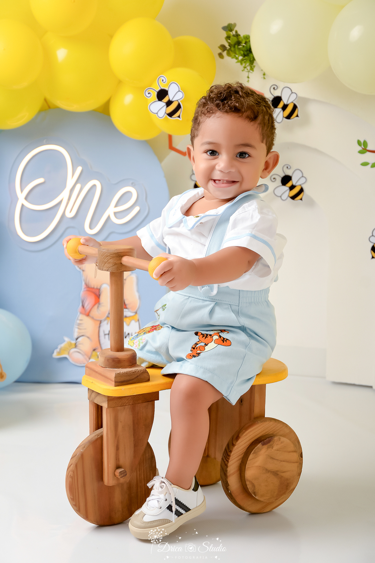 drica studio - smash the cake - 1 ano - aniversario - tema ursinho pooh - cenário com abelhas - one de led - painel azul - bebê sentado em motinha de madeira - bebê sorrindo 