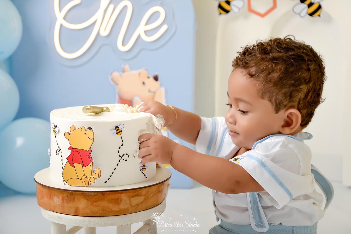 drica studio - smash the cake - 1 ano - aniversario - tema ursinho pooh - cenário com balões - abelhas - favo de mel - one de led - painel azul e branco - bolo branco do ursinho pooh - bebê tendo o primeiro contato com o bolo 