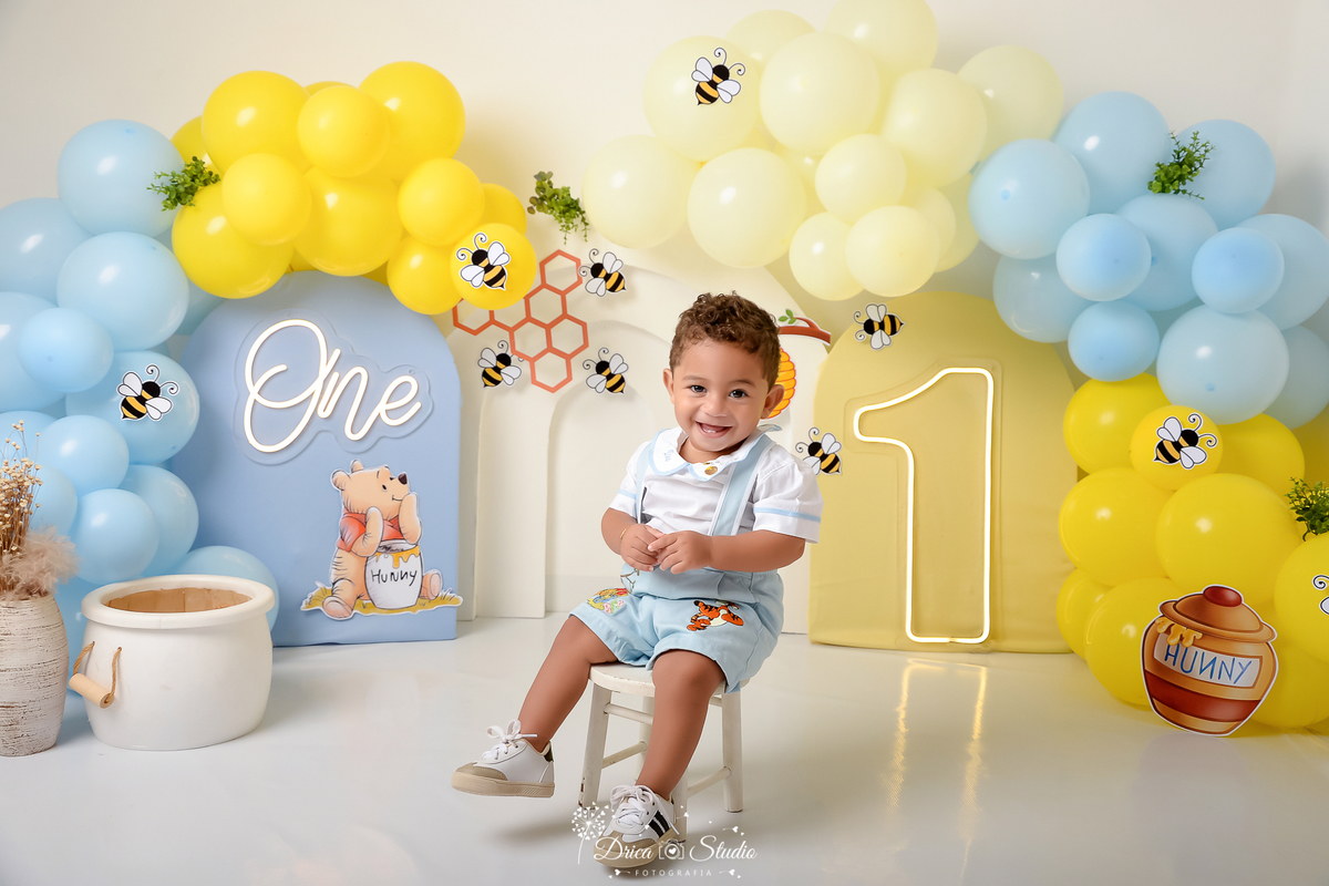 drica studio - smash the cake - 1 ano - aniversario  - ursinho pooh abelhas - favo de mel - cenário com balões - balões azuis e amarelos - one de lede - 1 de led - bebê sentado numa cadeirinha -  unsando uma roupinha personalizada do ursinho pooh